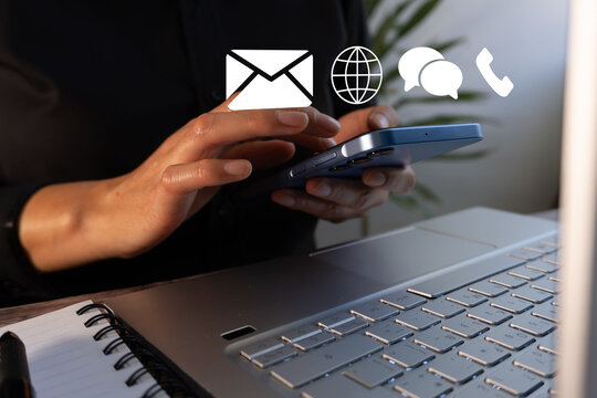 A woman uses a cell phone while sitting at a desk with a laptop. The laptop includes contact icons for a business: email, website, phone, and speech bubbles. - Powered by Adobe