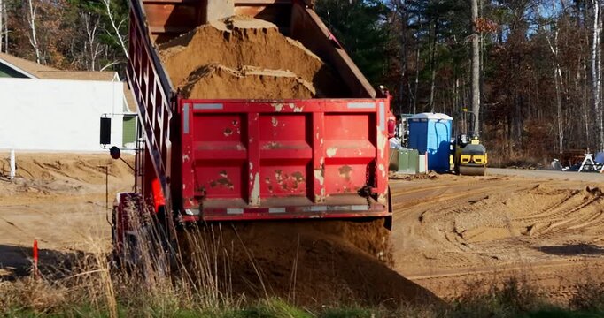 A load of brown dirt is emptied from the box of a red dump truck into a pile on a plot of real estate that will eventually contain a new home.