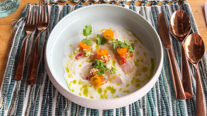 Slices of raw fish ceviche or crudo in a shallow bowl of white sauce, garnished with herbs, tomatoes, and olive oil