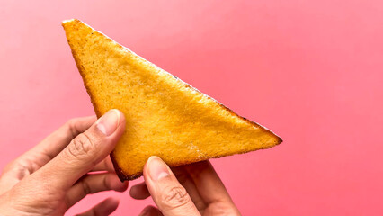 A triangular slice of golden-brown toasted bread, dusted with powder, held against a vibrant pink studio background