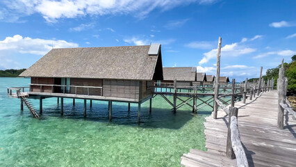 Several wooden overwater bungalows with pointed roofs stand on stilts, connected by a deck over transparent green-blue sea under a sunny sky
