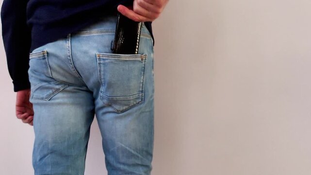 Hand demonstrates a wallet into a back pocket against a pale wall, focusing on the simple motion, the texture of fabric and leather, and the calm, uncluttered everyday setup