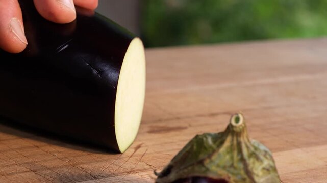 Slow-motion close-up of an eggplant being sliced into thin rounds in an outdoor kitchen. The knife moves smoothly through the purple flesh, revealing clean cuts and detailed vegetable texture.