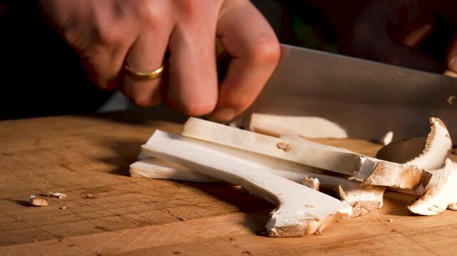 Close-up of king oyster mushrooms being sliced with a sharp knife on a wooden cutting board. The clean motion reveals the firm texture and smooth structure of the fresh mushrooms.
