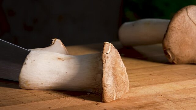 Close-up of a king oyster mushroom being cut cleanly in half with a sharp knife. The smooth motion reveals the dense texture and natural structure of the fresh mushroom.