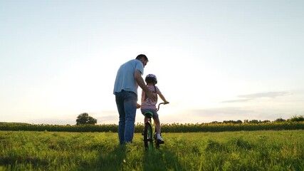 Loving father teaches toddler daughter training bicycle riding in sunset field. Father supports back of little girl riding bicycle in park. Father with daughter at bicycle riding lesson on holiday