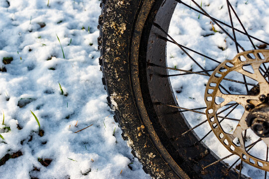 Close-up of a wide bike tire resting on patchy snow with emerging green grass tips underneath, wet and icy texture visible