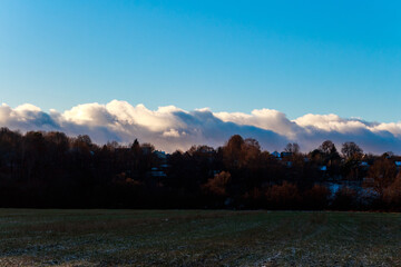 Autumn landscape with the first dusting of snow on a field, silhouetted dark trees topping a small hill beneath a dramatic, low band of bright clouds and vivid blue sky