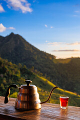 A gooseneck kettle and a shot glass of red liquid on a wooden deck, set against a stunning morning mountain landscape with golden hour sunlight and blue sky.