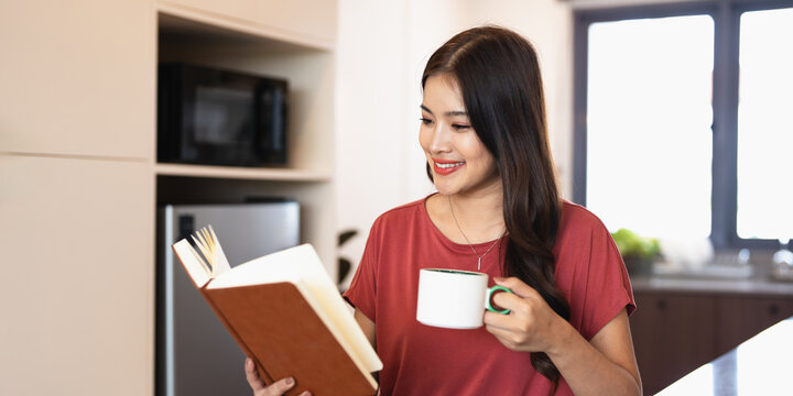 Pretty young woman with cup of coffee or tea reading book in modern kitchen counter at home apartment. Morning lifestyle concept. - Powered by Adobe