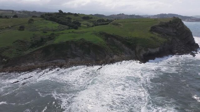 Side and upward drone shot on the Cantabrian coast, showing consecutive waves, bright green headlands, we see the town of Comillas in the distance, the sunlight reflecting softly on the sea.