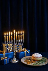 Hanukkah Jewish celebration vertical backdrop. Menorah with lit candles and gift boxes, a plate with a pastry, coins, and decor creating a warm mood . Dark blue background with space for text