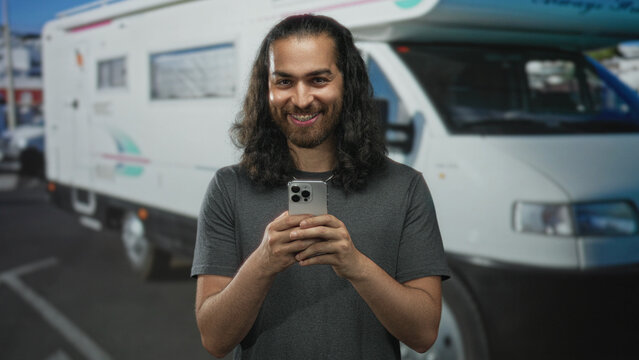 Young man holding smartphone and smiling in front of camper van on a city street while looking at screen; happiness travel. - Powered by Adobe