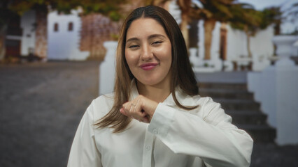 Woman in white shirt winking and punching with fist toward camera on sunlit historic street; confidence.