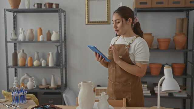 Woman wearing brown apron holds tablet and touches ceramic vase on wooden table in bright studio; craftsmanship passion. - Powered by Adobe