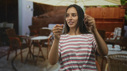 Woman smiling with hands held forward pinching gesture on street near restaurant terrace, fingers poised as if pulling thread; playful curiosity.