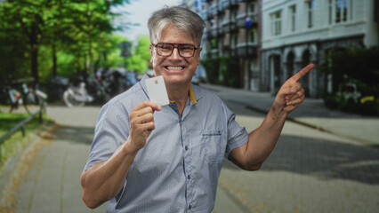 Fototapeta premium Man with grey hair holding a plain creditcard, smiling and pointing finger toward the right on a tree lined street; trust confidence promotion.