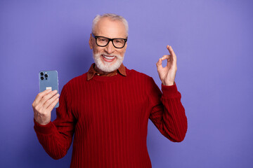 Happy senior man with smartphone wearing red sweater against purple background smiling for advertising