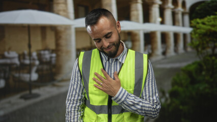 Young hispanic man wearing a reflective safety vest places his right hand on chest on a restaurant...