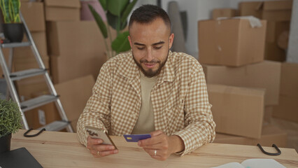 Man holding smartphone and creditcard with hands at table in building surrounded by moving boxes; new home contentment.