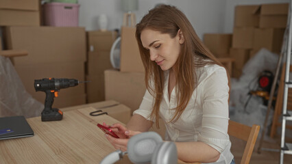 Woman taps smartphone on wooden table in building among moving boxes and power drill; calm preparation.