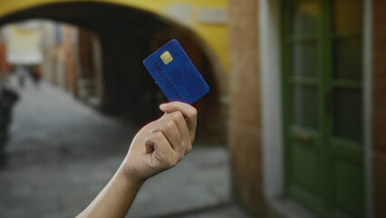 Hand of a caucasian man holding a blue credit card outdoors on a city street with green doors and...