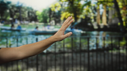 Man's hand raised against a blurred park backdrop with green trees and water indicating an outdoor garden setting on a sunny day.