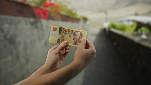 Man holding a 5000 forint banknote on a city street in hungary, with greenery and blurred urban background, showcasing outdoor urban life and currency handling.