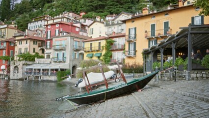 Fototapeta premium Blurred lakeside village scene with tilted moored boat, colorful facades, stone quay and terrace cafes in soft bokeh outdoor; background backdrop copyspace calm.