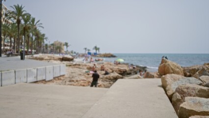 Obraz premium Coastal promenade defocused outdoor scene with soft shoreline, rocks and distant palms; background template copyspace calm.