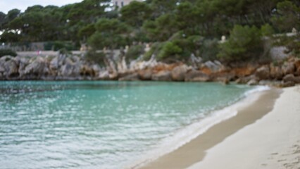 Blurred view of cala gat beach in mallorca featuring bokeh effect, defocused sandy shore, rocky coastline, and serene turquoise waters under a cloudy sky.