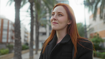 Woman with red hair in a city street setting looks thoughtful outdoors surrounded by palm trees wearing a black coat showcasing a serene urban environment.