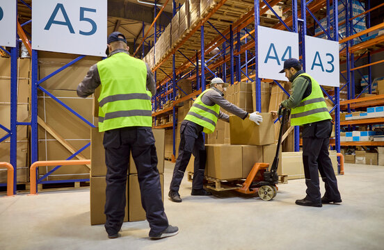 Warehouse workers inventory logistics teamwork boxes shipping. Team uses a pallet jack to move cartons, following aisle signage for storage and flow. Concept of efficient warehouse operations.