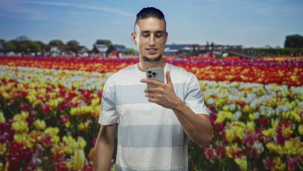 Fototapeta premium Man holding smartphone and looking down at the phone, hand visible over device against a studio tulip field backdrop; contemplation.