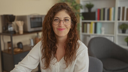 Woman with round glasses smiles at camera in an office with bookshelf and chair, showing freckled face and relaxed posture  calm confidence. © Krakenimages.com