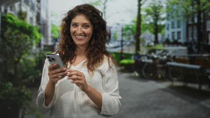 Woman holding smartphone and smiling while tapping the screen on a tree lined street by a canal building; digital connection joyful.