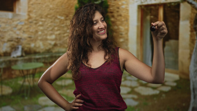 Young hispanic woman smiling, holding a cherry between fingers at building entrance with stone patio and doorway; home play joy.