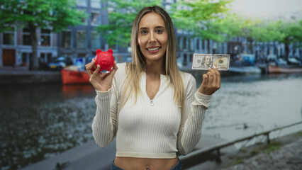 Young hispanic blonde woman holding a red piggy bank and a us banknote, smiling with hands raised...