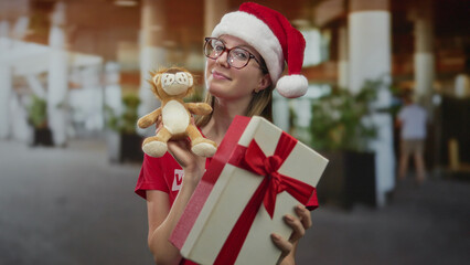 Woman wearing santa hat smiles and holds gift box and plush toy in building; holiday generosity...