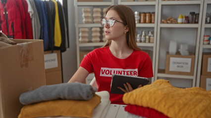 Obraz premium Volunteer woman inspects tablet while sorting clothing piles at table in building; compassion service solidarity.