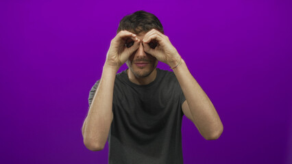 Young man looking through binocular gesture with both hands at purple studio wall; curiosity...