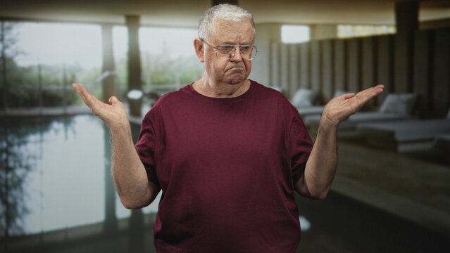 Senior man with glasses and maroon shirt shrugging with raised palms and pursed mouth beside an indoor pool and lounge chairs inside a building; confusion doubt.