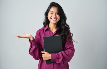 Young Asian woman holds notebook, smiles warmly, and gestures with open palm. She looks happy and engaged, ready to share ideas or information. This photo represents learning and presentation.