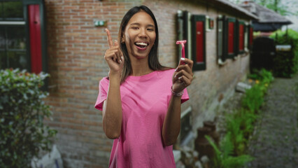 Thai woman holding pink razor and points index finger upward in front of building; self care confidence.
