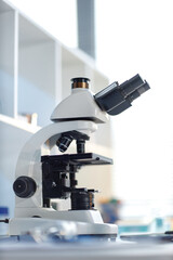 Laboratory microscope standing on table in scientific research setting, optical device featuring multiple objective lenses and adjustment knobs, used for biological or medical analysis