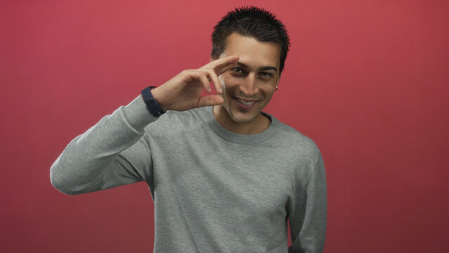 Hispanic man smiling confidently in front of an isolated red background wall, dressed in a casual grey sweater while posing with a friendly demeanor.