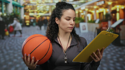 Woman holding basketball and clipboard, standing on busy street, wearing whistle, outdoor scene with shops and lights, focused expression, hispanic female coach.
