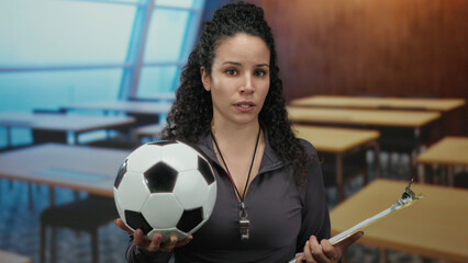 Hispanic woman holding soccer ball and clipboard in classroom with focused expression, teaching sports indoors in educational setting, wearing whistle and dark jacket