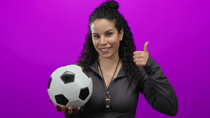 Young hispanic woman holding soccer ball and giving thumbs-up with a whistle around her neck against a vibrant pink background