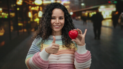 Young hispanic woman holding red heart-shaped object and gesturing thumbs up in brightly lit street with blurred background under night lighting.